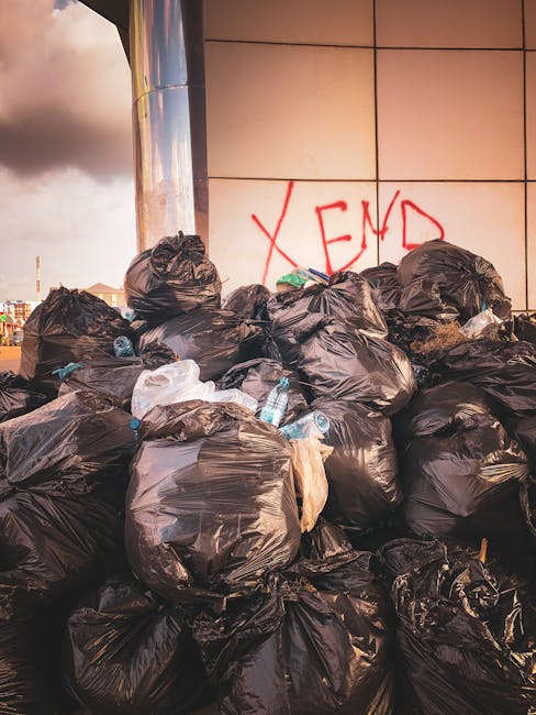 A weathered red metal dumpster with visible rust and peeling paint, situated against a dark green building wall and a light gray concrete surface in an urban environment. The dumpster is rectangular w