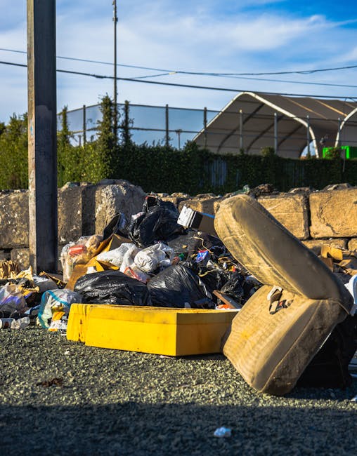 A large, weathered green waste dump container positioned on a paved urban area, filled with mixed rubbish including flattened cardboard boxes, smaller packing cartons, white plastic or paper packaging