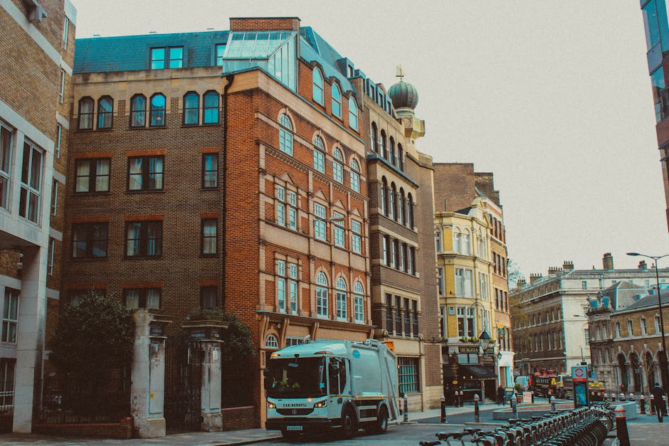The image shows a city street scene with several multi-story buildings featuring brick and ornate architectural details, typical of Central London's historic style. In the foreground, a white waste co