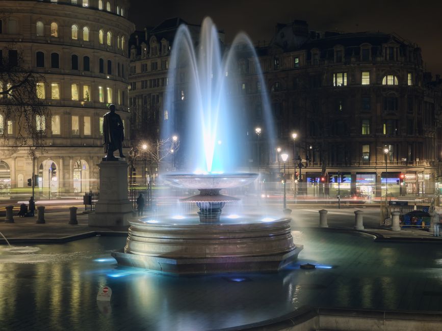 A night-time scene of Trafalgar Square featuring a circular stone fountain illuminated by blue lighting, with multiple jets of water rising from the center and cascading back into the basin. Surroundi